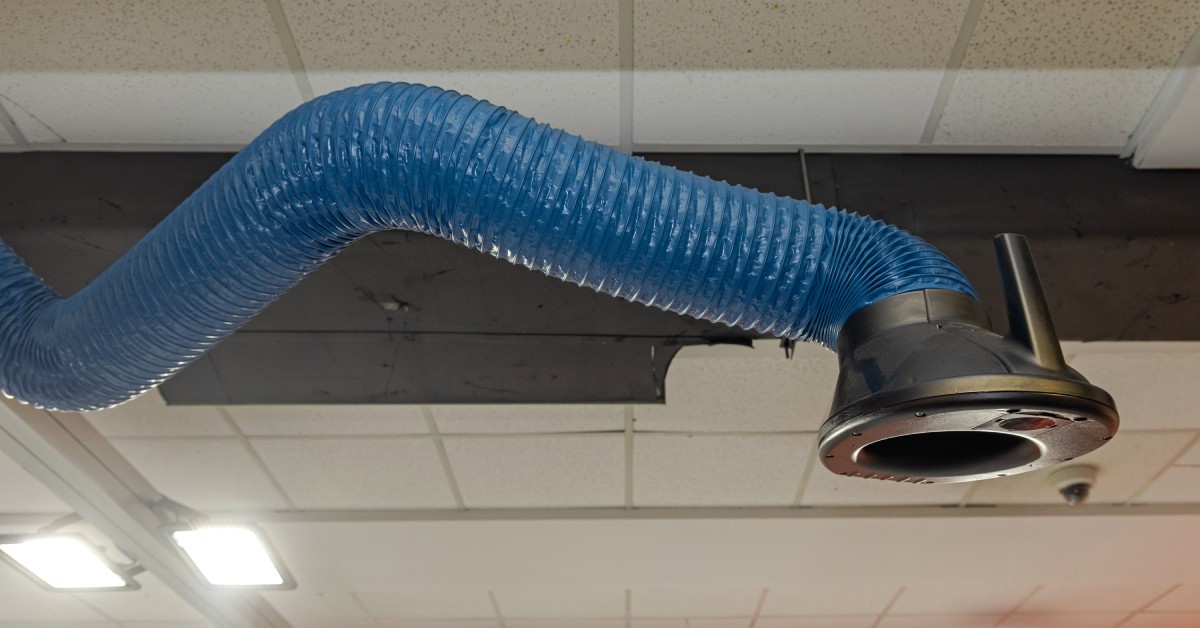 A welding fume extractor sits on the ceiling of a facility. It has an extended blue hose. Ceiling lights are nearby.
