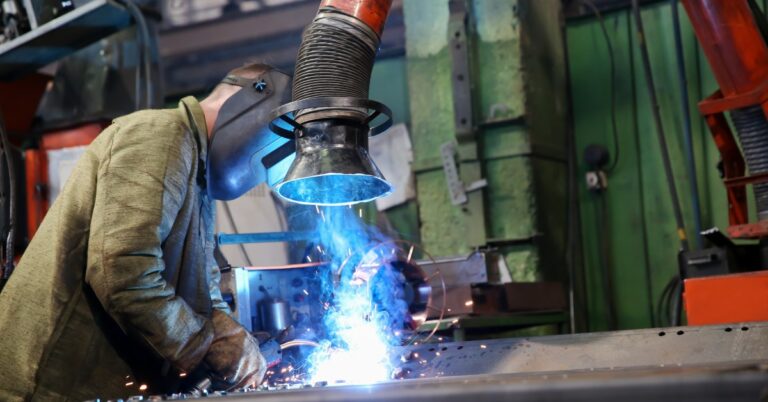 An employee welds in a factory while an extractor pulls the fumes out. The worker wears a welding mask.