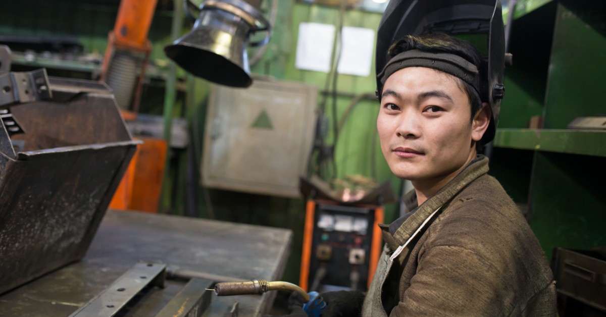 A worker prepares to weld in a factory, with his welding mask up. The extractor sits in the background.
