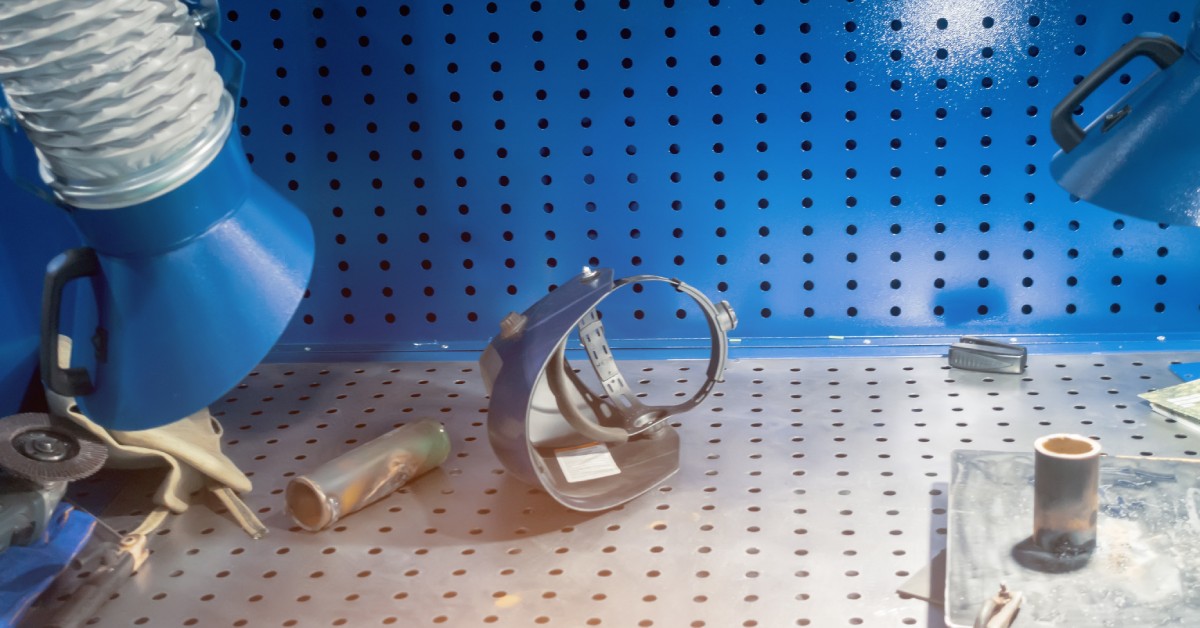A blue welding fume system tube hangs above a table that features various tools, including a welder's mask.