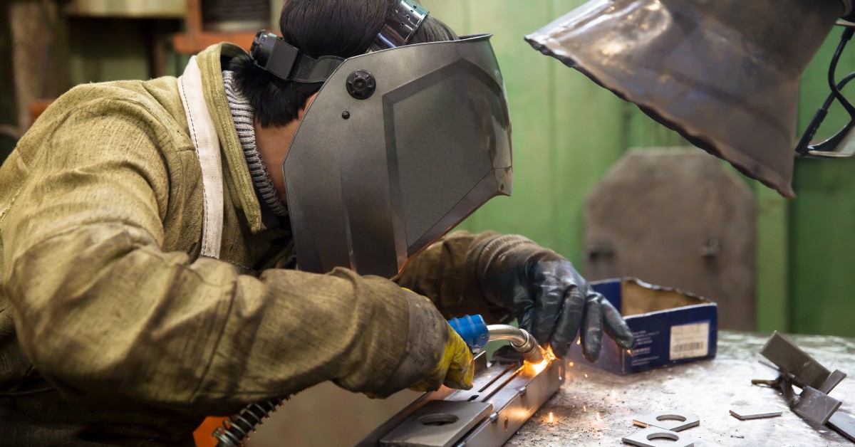 An industrial worker hunches over while welding a piece of metal. The front of a welding fume extractor hangs over them.