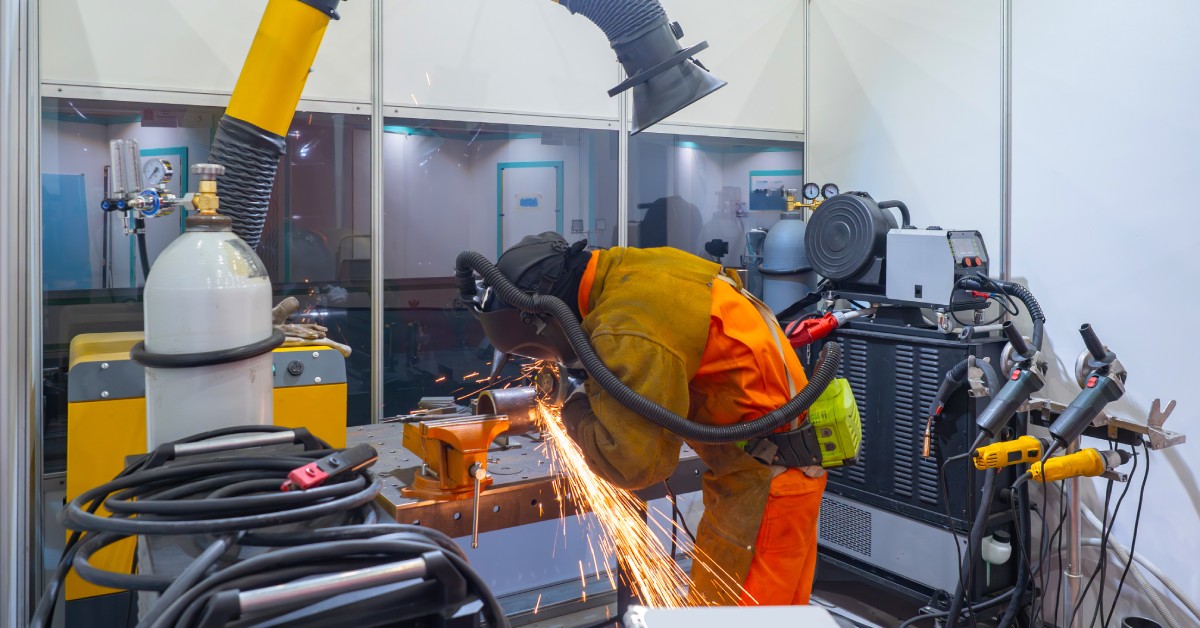 An employee leans down to work on a welding task while wearing protective equipment. A fume extractor sits nearby.