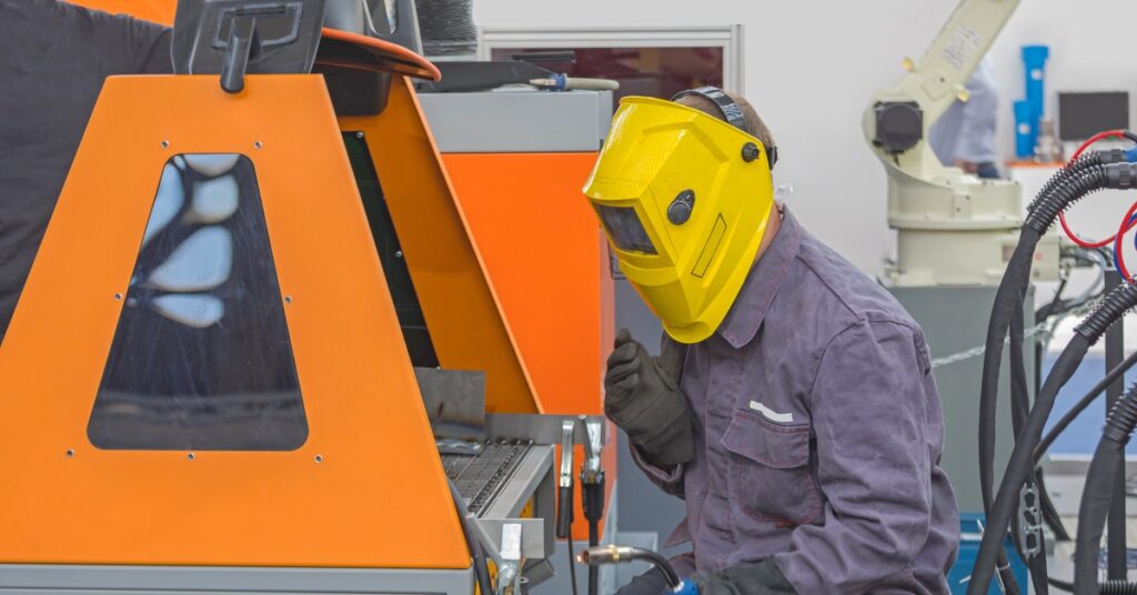 A worker wearing a jumpsuit, gloves, and a welding mask sits in front of an orange-walled welding booth.
