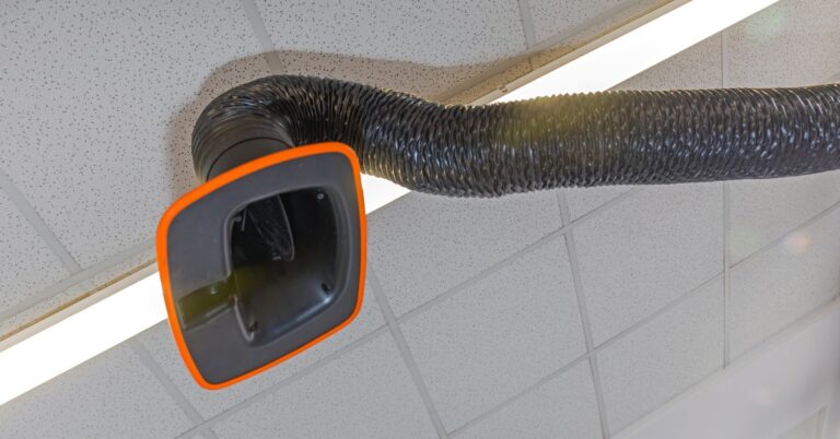 A welding exhaust arm extends across the ceiling of a shop. Its black tubing rests against the ceiling panels and light.