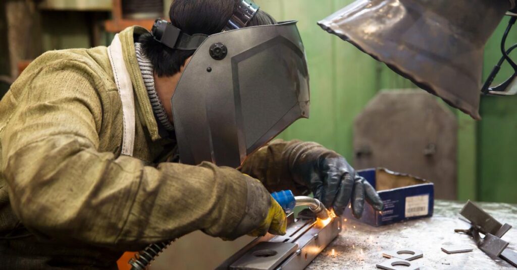 A worker wearing a black face mask welds a piece of metal on a table. An extractor hangs above him.