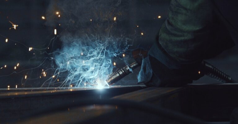 Close-up of a worker's gloved hands as they perform welding. Smoke rises up from the point of contact.