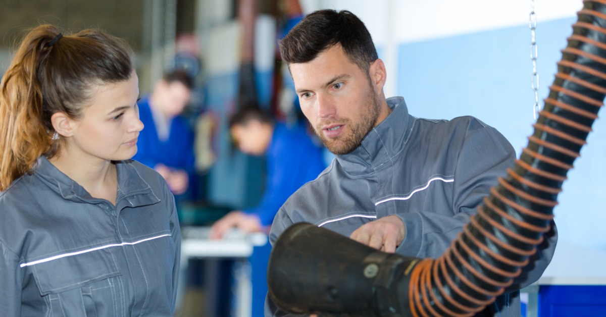 A worker in a jumpsuit shows another employee a fume extractor. The two stand together in a workshop.